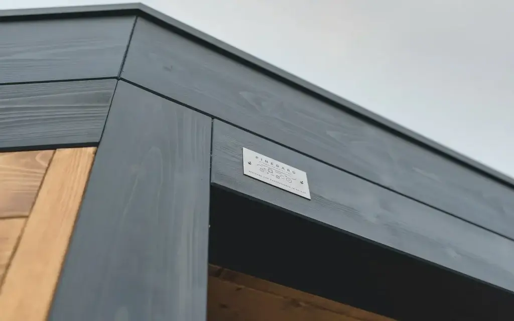 Auf dem Bild sieht man eine Nahaufnahme der Ecke einer modernen Holzpergola in dunkler Farbe. An der Konstruktion befindet sich ein kleines Metallschild mit dem Logo „PINEGARD“.