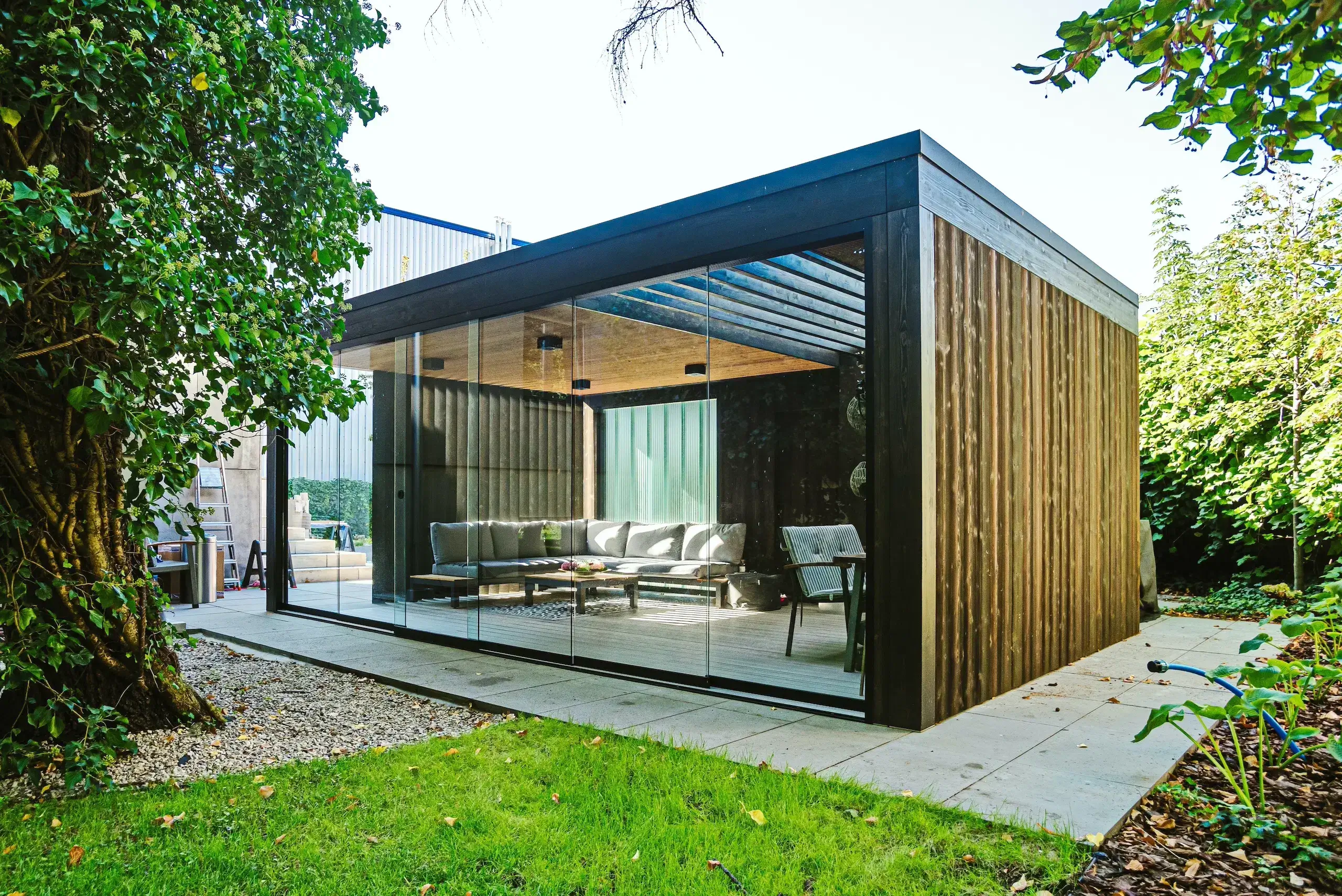 Das Bild zeigt einen Moderne Gartenhäuser. Es hat große Glaswände vom Boden bis zur Decke. Draußen sind Bäume, Gras und Sträucher zu sehen. Die Außenwände bestehen aus vertikalen Holzpaneelen und einem dunklen Rahmen. Innen stehen eine Eckcouch, ein Tisch und ein Sessel. Die Decke ist aus Holz und darauf sind Lampen angebracht. Alles steht auf einer befestigten Terrasse und wirkt gemütlich.