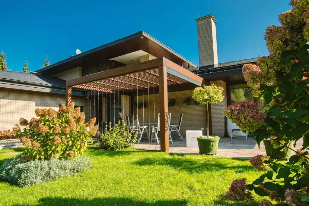 Auf dem Bild sieht man einen gepflegten Garten mit einer modernen Terrasse und einer großen Holzpergola am Haus. Unter der Pergola stehen Gartenmöbel mit Tisch und Stühlen, die einen gemütlichen Sitzbereich bilden. Umgeben ist die Terrasse von grünem Rasen, Sträuchern und blühenden Pflanzen, während das Haus mit heller Fassade und großem Kamin im Hintergrund zu sehen ist.