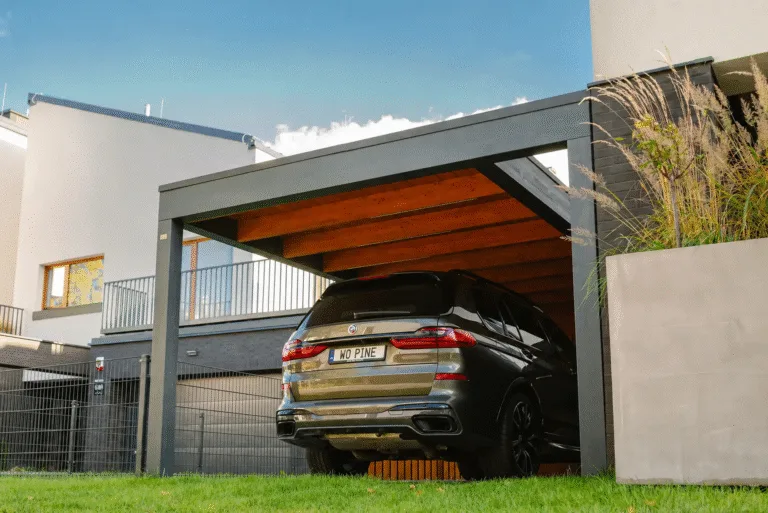 Das Foto zeigt einen modernen Wohnbereich mit einem eleganten Carport, unter dem ein schwarzes Auto geparkt ist. Die Konstruktion des Carports verbindet die Holzdecke mit dunklen Metallpfosten und schafft ein minimalistisches und funktionales Design.