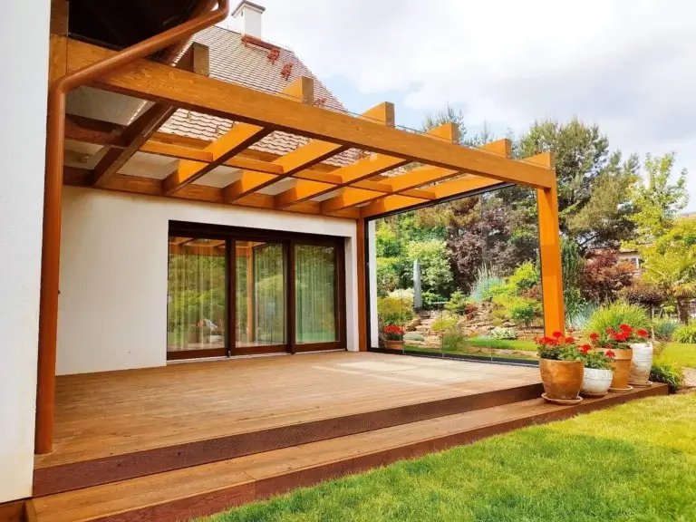 Die Grafik zeigt eine moderne Terrasse am Haus. Sie hat einen Holzboden und zwei Stufen, die auf den Rasen führen. Über der Terrasse befindet sich ein Pergola mit transparentem Dach. Große, schiebbare Glastüren führen ins Innere des Hauses. Auf der Terrasse stehen Blumentöpfe mit roten Blumen. Im Hintergrund ist ein Garten mit Sträuchern, Bäumen und Blumen zu sehen.