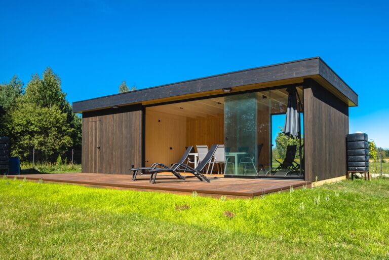 Die Grafik zeigt eine moderne Gartenpergola aus Holz mit dunklen Holzwänden und einem flachen Dach. Vorne befinden sich große, gläserne Schiebetüren. Vor dem Eingang gibt es eine Holzterrasse mit Sesseln und einem Tisch. Im Inneren sind helle Holzpaneele zu sehen. Neben dem Häuschen stehen schwarze, runde Behälter. Das Ganze ist von Gras und Bäumen umgeben – ein friedlicher Ort zum Entspannen.