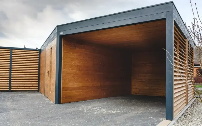 Auf dem Foto sehen wir einen modernen Holz Carport. Die Konstruktion ruht auf einem dunklen Gestell, und die Wände bestehen aus horizontalen Holzpaneelen. Auf der rechten Seite befindet sich ein kleiner Bereich mit Kies und einem Baum.