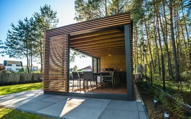 Eine moderne Gartenlaube mit Holz- und Metallkonstruktion schafft einen eleganten Essbereich im Freien. Im Inneren befinden sich ein Tisch mit Stühlen sowie ein eingebauter Grill, und alles wird von Lamellenwänden und einem Holzfußboden umgeben. Die Umgebung mit Steinplatten, Rasen und Grün unterstreicht den geordneten, einladenden Charakter des Bereichs — ideal für Treffen und zum Entspannen im Garten.