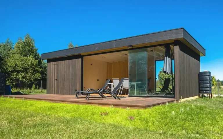 Auf dem Foto ist ein modernes hölzernes Gartenhaus mit Flachdach und dunkler Holzfassade zu sehen. Glastüren führen auf eine Holzterrasse, auf der schwarze Liegestühle sowie ein kleiner Tisch mit Stühlen stehen. Die gesamte Szenerie ist von grünem Gras und Bäumen umgeben. Rechts sieht man einen Stapel zylindrischer Behälter, wahrscheinlich zur Regenwassersammlung.