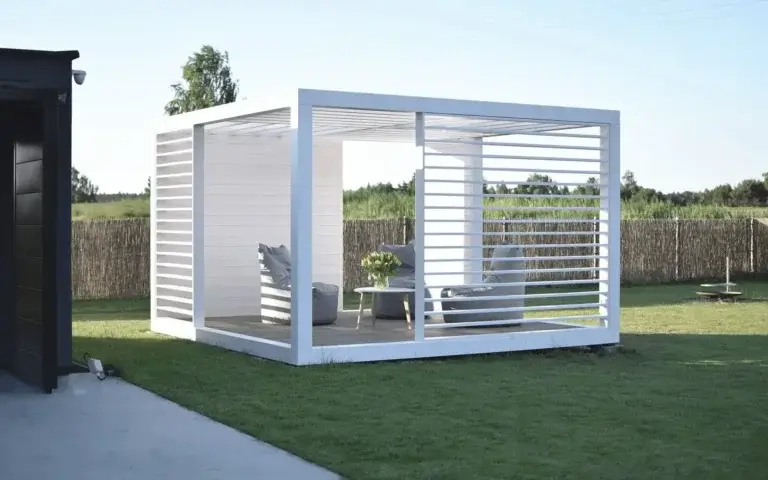 Auf dem Foto sehen wir eine moderne Holzpergola aus weißen, horizontalen Lamellen. Die Konstruktion sorgt für Halbschatten und lässt gleichzeitig Licht und Luft durch. Innen befinden sich drei bequeme Sessel mit Kissen sowie ein weißer Tisch mit einem Blumenstrauß. Die Pergola steht auf einem grünen Rasen, im Hintergrund ist eine natürliche Umzäunung aus vertikalen Holzstäben sowie ein Teil eines dunklen Gebäudes zu sehen.