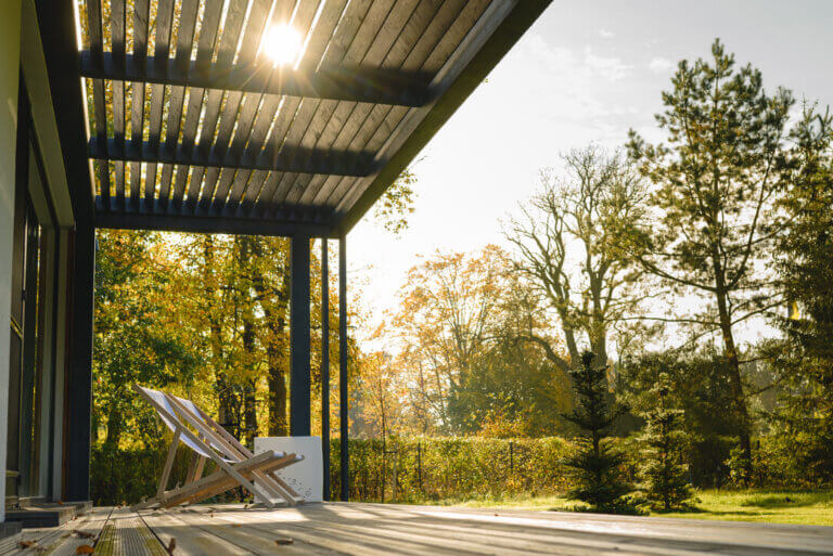 Das Foto zeigt eine ruhige Erholungszone auf einer Terrasse mit Holzboden und moderner Pergola, durch deren Lamellen warmes Sonnenlicht fällt und dekorative Schatten erzeugt. Auf der Terrasse stehen zwei Holzliegestühle, die zum üppigen Garten mit vielen Bäumen und Grünpflanzen ausgerichtet sind.