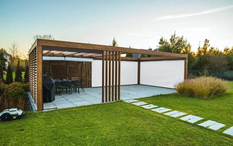 Das Bild zeigt eine moderne Gartenpergola mit einer Holzkonstruktion und vertikalen Lamellen an den Seiten, die teilweisen Schatten und Privatsphäre bieten. Im Inneren befinden sich ein Tisch mit Stühlen sowie ein Grill, was darauf hindeutet, dass der Raum als Ort für gemeinsames Beisammensein und Entspannung genutzt wird. Auf dem Boden sind große quadratische Platten verlegt, und an den Seiten sind weiße, aufrollbare Stoffplanen montiert. Die Umgebung wird durch einen gepflegten Rasen mit Steinplatten, die zur Pergola führen, gestaltet.