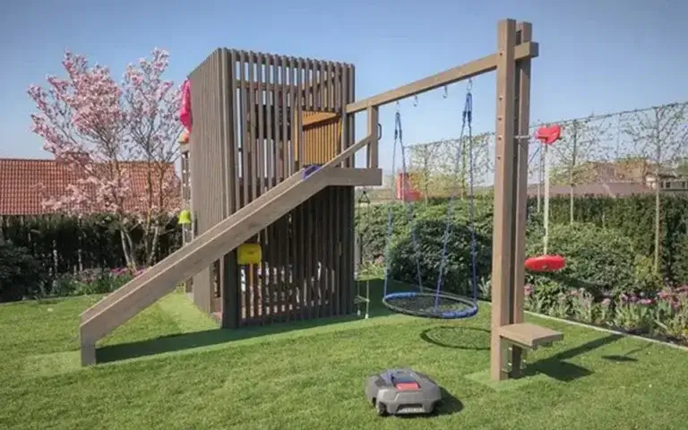 Das Foto zeigt einen modernen Spielplatz im Garten, der hauptsächlich aus Holz gefertigt ist. Er besteht aus einem Kletterturm mit vertikalen Lamellen, einer Rutsche sowie einer Schaukelanlage, darunter ein rundes Netz und eine rote Scheibe. Alles ist auf einem gepflegten Rasen angelegt, umgeben von Blumenbeeten und Bäumen.