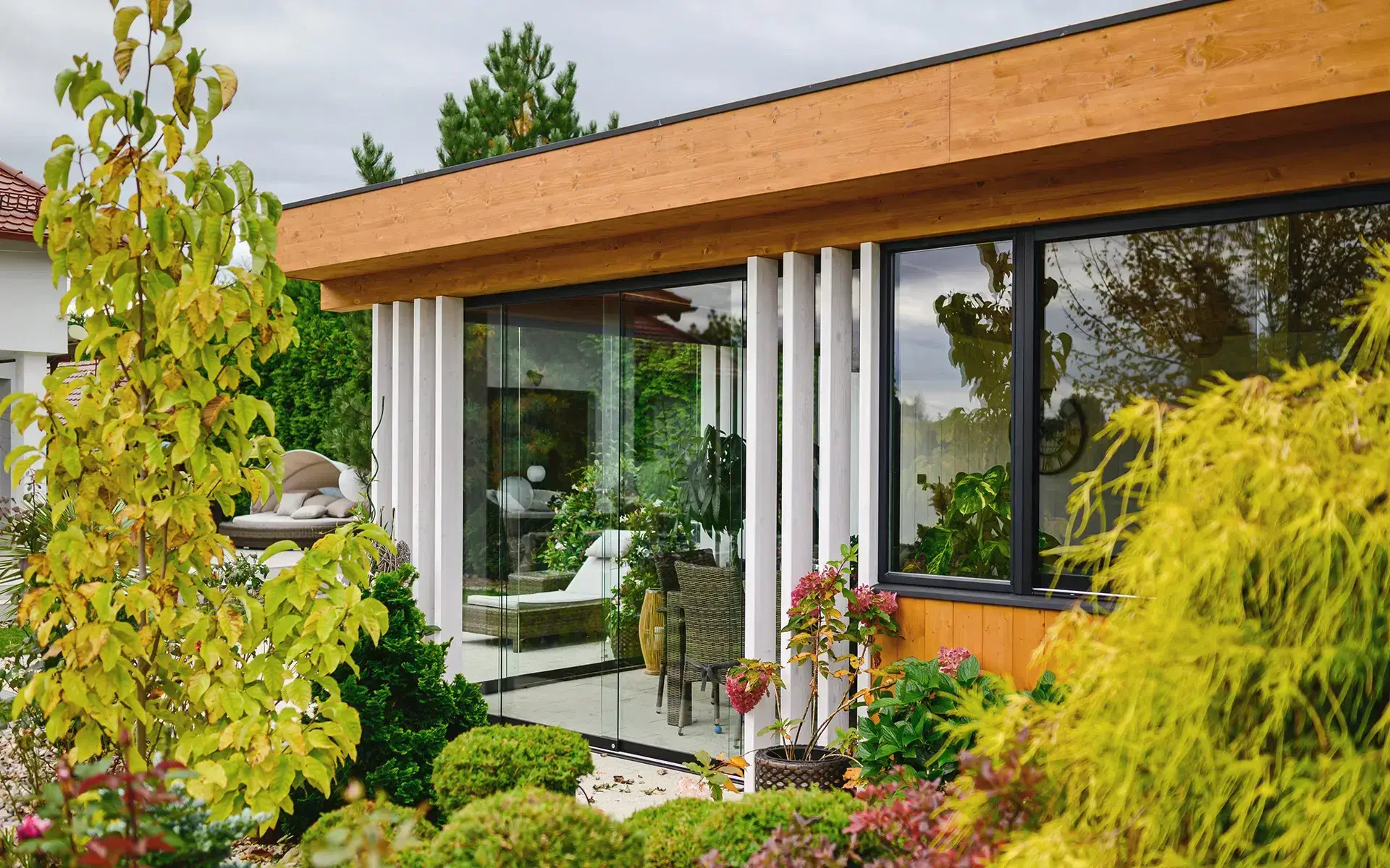 Das Foto zeigt ein modernes Haus mit einer Fassade aus hellem Holz, großen Glasfronten und einem flachen Dach mit einem hölzernen Dachüberstand. Ein charakteristisches Merkmal sind die vertikalen weißen Lamellen. Durch die Glaswände sind Korbmöbel und Pflanzen zu sehen, was die gemütliche, natürliche Atmosphäre des Innenraums unterstreicht. Der umgebende Garten ist üppig und gepflegt, voller Sträucher, Bäume und Grünflächen, und schafft einen harmonischen Raum zum Leben in der Nähe der Natur.